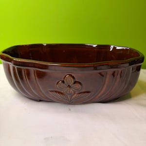 May include: A brown ceramic planter with a glossy finish. The oval-shaped planter features a ribbed design and a floral emblem on the front. The planter is suitable for indoor or outdoor use.