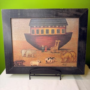 May include: A framed print of Noah's Ark, featuring a red ark with multiple windows and a black frame. The image includes animals such as elephants, giraffes, and camels. The print is displayed on a white surface.