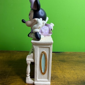 Peut inclure: Une figurine en c&eacute;ramique de Minnie Mouse posant son menton sur sa main tout en s'appuyant sur un piano droit de couleur cr&egrave;me. Elle porte une robe violette &agrave; pois blancs et un n&oelig;ud assorti. Le piano pr&eacute;sente des d&eacute;tails ovales dor&eacute;s.