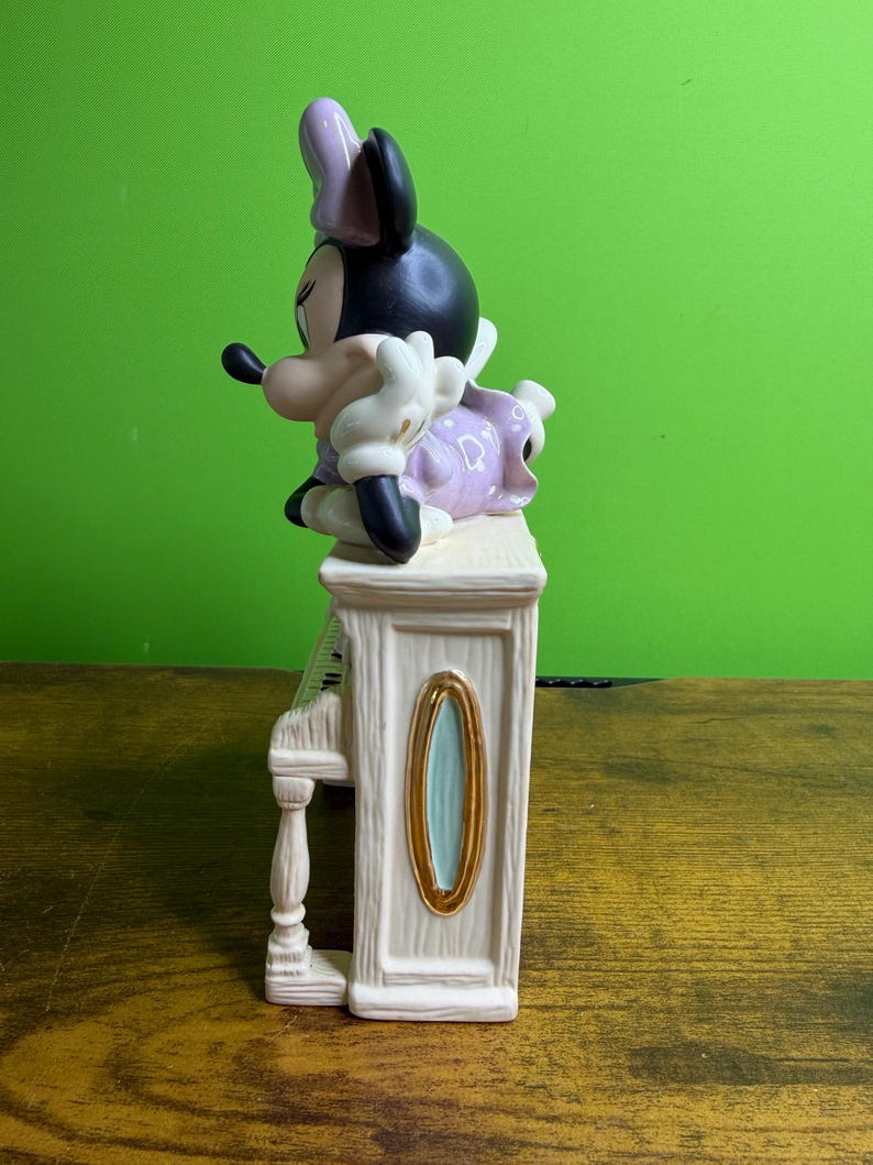 Peut inclure: Une figurine en c&eacute;ramique de Minnie Mouse posant son menton sur sa main tout en s'appuyant sur un piano droit de couleur cr&egrave;me. Elle porte une robe violette &agrave; pois blancs et un n&oelig;ud assorti. Le piano pr&eacute;sente des d&eacute;tails ovales dor&eacute;s.