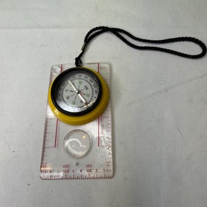 May include: A yellow and black compass with a clear ruler base and a black lanyard. The compass has a clear magnifying glass and measurement markings in inches. The compass needle points north.