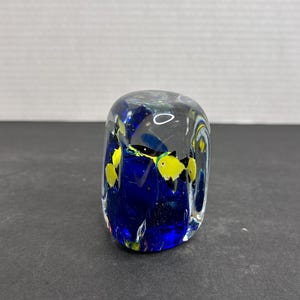May include: A clear glass paperweight with a blue and yellow design. The paperweight features a scene of fish swimming in a blue ocean.