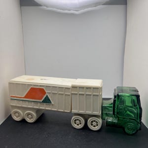May include: A vintage toy truck with a green cab and a white trailer. The trailer has a red and green geometric design. The truck has white wheels and is set against a dark background.