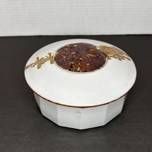 May include: A white ceramic trinket box with a round lid. The lid has a brown and gold design with a circular pattern in the center.