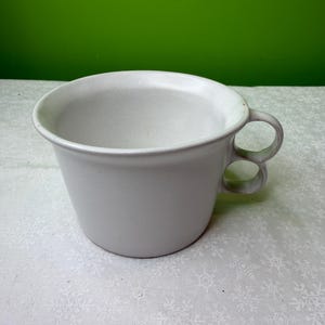 Può includere: Una tazza in ceramica grigio chiaro con un design unico del manico. Il manico presenta due fori per le dita. La tazza ha un'ampia apertura e un bordo leggermente svasato. Lo sfondo è di colore verde.