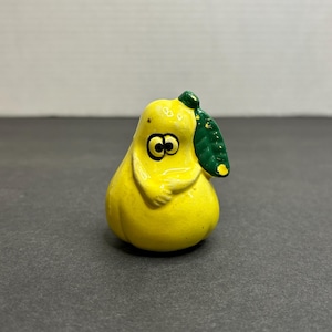 Puede incluir: Una figura caprichosa en forma de pera amarilla, con ojos y brazos de dibujos animados. Tiene una hoja y un tallo verdes. La pera está pintada con un acabado brillante, y el diseño es juguetón y único.