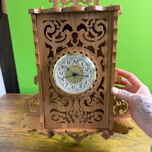 May include: A wooden clock with an ornate, carved design. The clock face is round with black numerals and hands. The clock is housed in a rectangular wooden frame with decorative cutouts. The clock is approximately 10 inches tall.