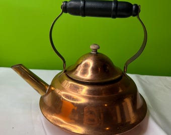 May include: A copper-colored tea kettle with a black wooden handle and a small wooden knob on the lid. The kettle has a curved spout and a shiny, reflective surface. The handle is attached to the kettle with brass-colored metal.