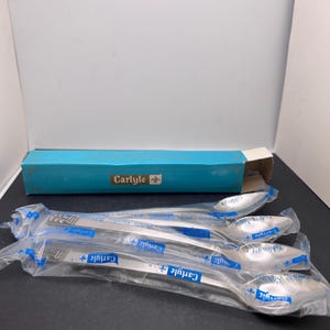 Könnte beinhalten: Ein Set aus vier Carlyle Edelstahl-Löffeln, einzeln in Klarsichtfolie verpackt. Die Löffel sind silberfarben mit einem dekorativen Griffdesign. Im Hintergrund ist eine türkisfarbene Schachtel mit dem Carlyle-Logo zu sehen.