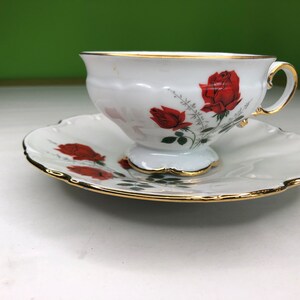 Puede incluir: Juego de taza y platillo blancos con diseño de rosas. La taza y el platillo tienen un borde dorado. La taza presenta un diseño de rosas rojas con hojas verdes. El platillo tiene un borde festoneado y un diseño de rosas a juego.