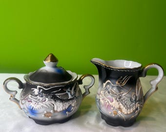 Op de afbeelding: Een zwarte en witte porseleinen roomkan en suikerpot set met gouden accenten. Beide stukken hebben een gedetailleerd drakenontwerp.