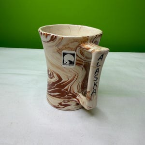 May include: A ceramic mug with a marbled brown and cream design. The handle is inscribed with the word "ALASKA" in black letters. A small sticker with a bear silhouette is visible on the mug.