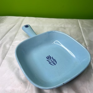 Può includere: Un piatto da portata quadrato azzurro con manico. Il piatto presenta un motivo floreale blu scuro al centro. Il manico ha un piccolo disegno blu coordinato. Il piatto è in ceramica o smalto.
