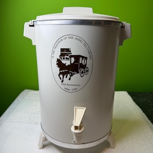 May include: A vintage white Jewel Tea Company coffee urn with a brown logo featuring a horse-drawn carriage. The urn has a spigot, a lid, and a handle on each side. The logo commemorates the 95th anniversary, 1899-1994.