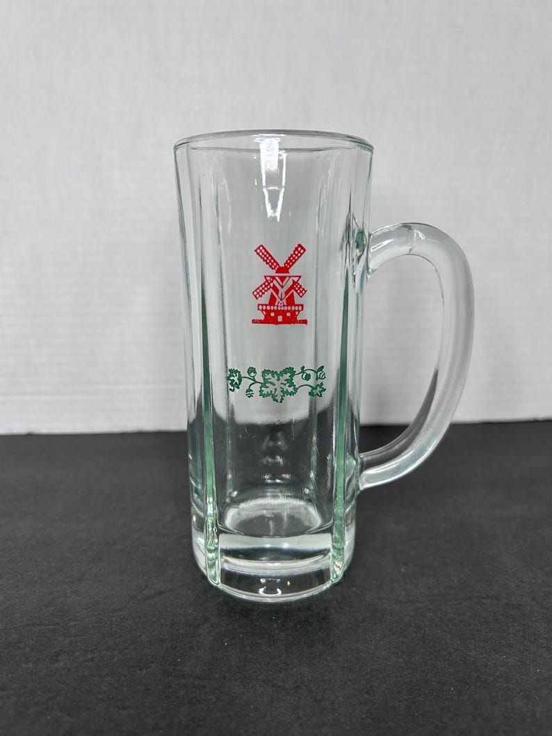 May include: A clear glass beer mug with a handle. The mug has a red windmill design with green vines around the bottom.