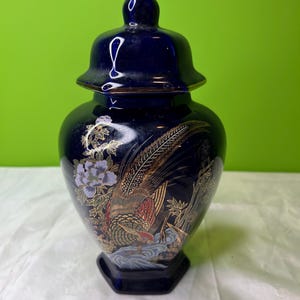 May include: A dark blue ceramic jar with a lid, featuring a detailed painting of a pheasant and floral designs in gold, red, and purple. The jar has a hexagonal base and a glossy finish.