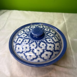 Puede incluir: Un cuenco de cerámica azul y blanco con tapa. El cuenco tiene forma redondeada con borde azul y un estampado floral blanco en la tapa. La tapa tiene un pequeño asa redonda azul.