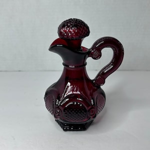 Op de afbeelding: Een robijnrode glazen fles met een getextureerd oppervlak en een bijpassende stop. De fles heeft een handvat en een rond lichaam.