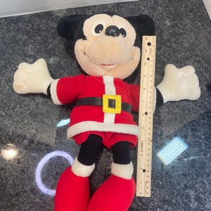 May include: A plush Mickey Mouse toy wearing a red and white Santa suit with a black belt and yellow buckle. The toy is positioned next to a wooden ruler, showing a total height of approximately 12 inches from the top of the head to the feet.