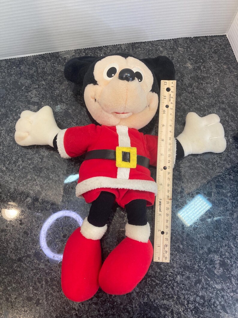 May include: A plush Mickey Mouse toy wearing a red and white Santa suit with a black belt and yellow buckle. The toy is positioned next to a wooden ruler, showing a total height of approximately 12 inches from the top of the head to the feet.