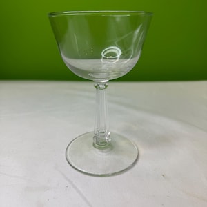 Puede incluir: Una copa de cóctel transparente con un cuenco ancho y poco profundo, una base redonda y un tallo delgado y acanalado. El vaso está sobre un fondo verde. Ideal para cócteles.