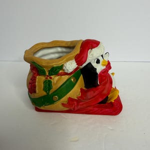 May include: A ceramic Christmas decoration featuring a penguin wearing a Santa hat and red scarf, peeking out of a yellow sack. The sack is decorated with green holly leaves and a red ribbon.