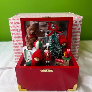 Puede incluir: Una caja de exhibición navideña de madera roja con frente transparente, con figuras en miniatura. En el interior hay un oso de peluche, un árbol de Navidad, un cascanueces y figuras con sombreros rojos. La caja tiene esquinas doradas.