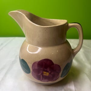 Puede incluir: Jarra de cerámica color beige con cuerpo redondo y pico curvo. La jarra está decorada con una gran flor de color rojo oscuro y hojas verdes. Tiene un asa y es adecuada para servir bebidas.