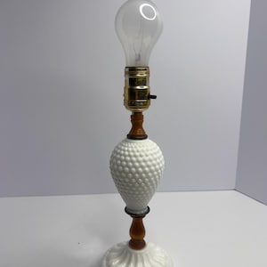 May include: A white hobnail table lamp with a clear light bulb. The lamp has a brass-coloured socket, a wooden stem, and a round, ridged base. The lamp is turned off.