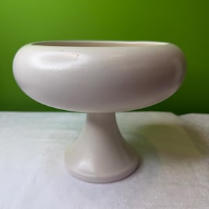 Puede incluir: Un cuenco de pedestal de cerámica blanca, con una parte superior redondeada y una base acampanada. El cuenco es de color blanco roto y se asienta sobre una superficie blanca sobre un fondo verde. El cuenco probablemente se utiliza con fines decorativos.