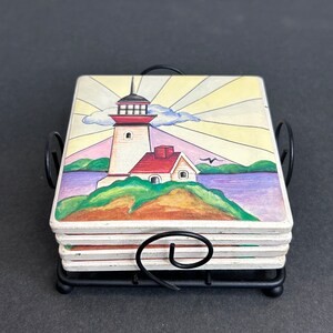 May include: Set of four ceramic coasters featuring a lighthouse with a red roof and a white house on a green island. The coasters are in a black metal holder with a sunburst design in the background.