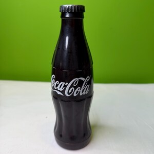 May include: A dark brown Coca-Cola bottle with a silver cap and white Coca-Cola logo. The bottle has a classic, contoured shape and is set against a green background.
