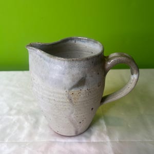 Può includere: Un'anfora in ceramica bianca con una glassa macchiata e un manico marrone. L'anfora ha una forma leggermente arrotondata e una bocca larga.