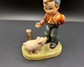 May include: A porcelain figurine of a boy wearing a blue hat with stars, holding a ball, and looking at a small pink pig. The figurine is standing on a green base with a brown wooden post.