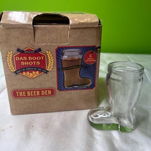 May include: A clear glass boot-shaped shot glass stands beside a cardboard box. The box features the text "Das Boot Shots" and "The Beer Den". A sticker on the box says "6 Shot Glasses". The shot glass is empty.