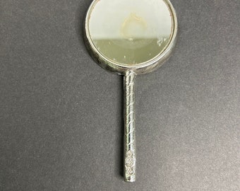 Könnte beinhalten: Ein silberner Handspiegel mit einer runden, reflektierenden Oberfläche und einem gedrehten Griff.