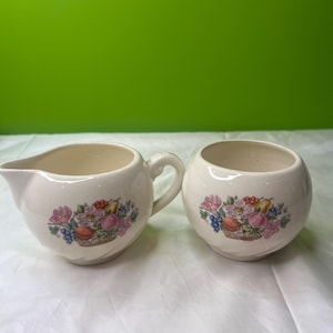 Könnte beinhalten: Cremé-farbenes Sahne- und Zuckerdosen-Set. Jedes Stück hat ein Blumen- und Obstkorb-Design in Rosa, Lila, Blau und Gelb. Der Sahneaufgießer hat einen Griff und einen Ausguss, während die Zuckerdose rund ist.