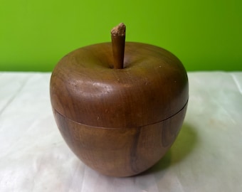 Könnte beinhalten: Eine holzfarbene, apfelförmige Box mit einem Stiel. Die Box besteht aus dunkelbraunem Holz und hat eine glatte Oberfläche. Der Stiel ist hellbraun und scheint aus einer anderen Holzart zu bestehen. Die Box steht auf einer weißen Oberfläche mit grünem Hintergrund.