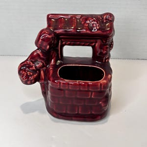May include: A small, red ceramic planter shaped like a brick well with a woman figure on the side. The planter has a small opening at the top for planting.