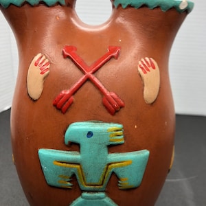 Peut inclure: Un vase en poterie de terre cuite avec un motif turquoise et brun. Le vase a deux becs verseurs et une anse. Le motif comprend un oiseau stylisé, des flèches croisées et des empreintes de mains.