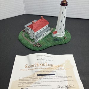 May include: A certificate of authenticity for a Danbury Mint Sandy Hook Lighthouse sculpture. The certificate states that the sculpture is a re-creation of the historic lighthouse, sculpted with exquisite detail and painted by hand in true-to-life colors. The serial number is AA1702.