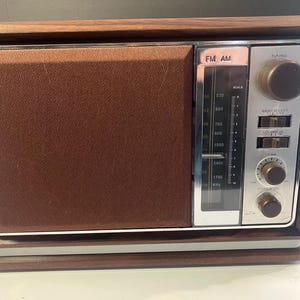 May include: A vintage FM/AM radio in a brown and silver finish with a wooden frame. The radio has a brown speaker cover, a silver control panel with tuning dials, and a frequency scale. A retro style electronic device.