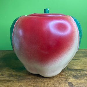 May include: A ceramic apple-shaped canister with a removable lid. The body features a gradient of vibrant red transitioning to a creamy off-white at the base. A small green stem sits atop the lid, and two green leaves are visible on the sides.