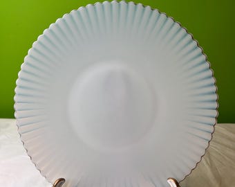 Könnte beinhalten: Ein weißer Milchglas-Dinnerteller mit einem gewellten Rand und einem blau-weißen, irisierenden Finish. Der Teller steht auf einem goldenen Metallständer.