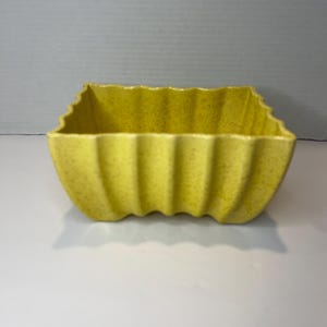 May include: A yellow ceramic planter with a square shape and a rippled design. The planter has a speckled texture.