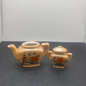 Op de afbeelding: Een miniatuur keramische theeset, bestaande uit een theepot en een suikerpot. De set is perzik van kleur met handgeschilderde bloemen- en pagodepatronen. De theepot heeft een tuit en een handvat, en de suikerpot heeft een deksel en twee handvatten.