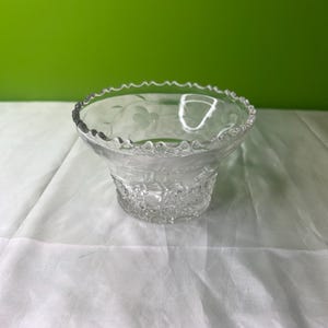 Puede incluir: Un tazón de vidrio transparente con un borde festoneado y un diseño floral grabado. El tazón está sobre una superficie blanca.