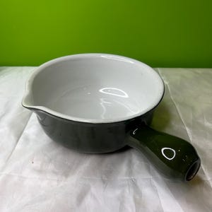 May include: A dark green enamel saucepan with a white interior and a spout. The saucepan has a single, rounded handle and a black rim. The background is a solid green color.