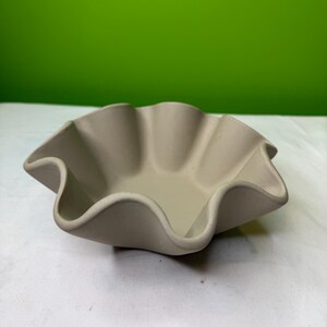 Op de afbeelding: Een beige keramische kom met een gerimpelde rand. De kom heeft een golvend, geschulpt ontwerp rond de rand. De kom is leeg en staat op een wit oppervlak met een groene achtergrond.