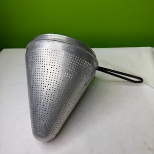May include: A silver-colored, cone-shaped kitchen strainer with numerous small holes. It has a black handle and is designed for straining liquids or rinsing food items. The strainer is made of metal and sits on a white surface.